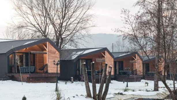 A row of cabins with wooden exteriors and large windows, situated in a snowy landscape with a calm body of water in the foreground.