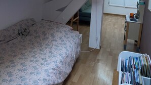 Une petite pièce avec un plafond en pente, un lit recouvert d’une courtepointe à motifs floraux, un bureau avec une chaise et une bibliothèque.