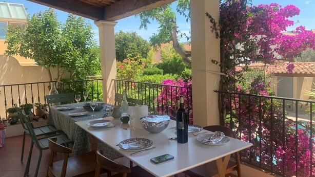 Un espace extérieur abrité, aménagé pour les repas, avec une table dressée pour quatre personnes, entouré de plantes en pot et offrant une vue sur un jardin aux fleurs roses.