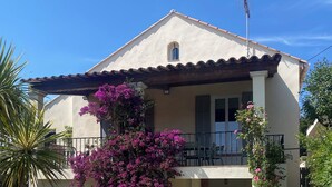 Une maison à deux étages avec une piscine, un balcon agrémenté d’une pergola et un jardin où poussent des bougainvilliers aux fleurs violettes.