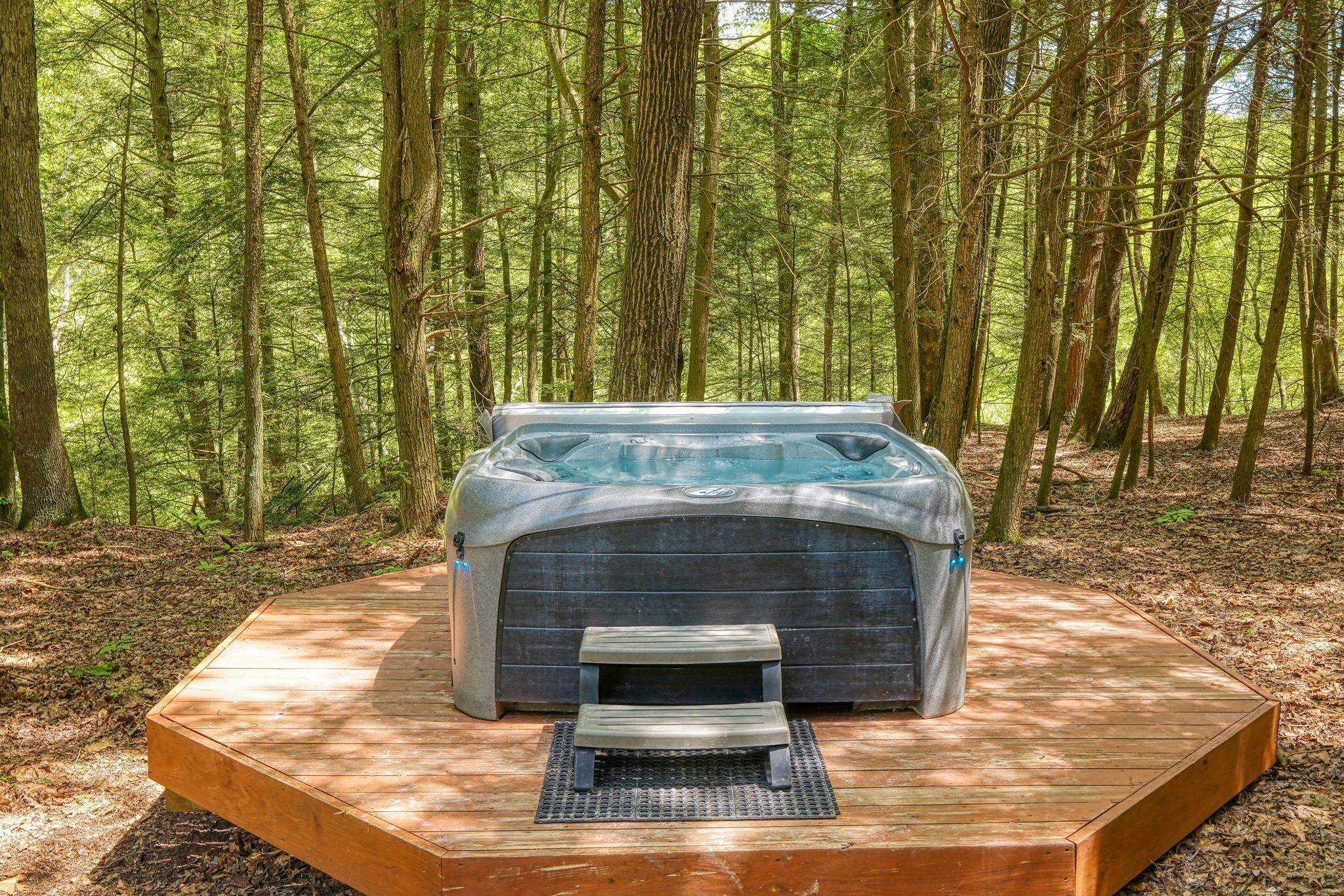 A hot tub in a forest setting with a wooden deck and a small bench.