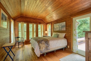 A wooden cabin bedroom with a bed, a chair, a table, and a painting on the wall.