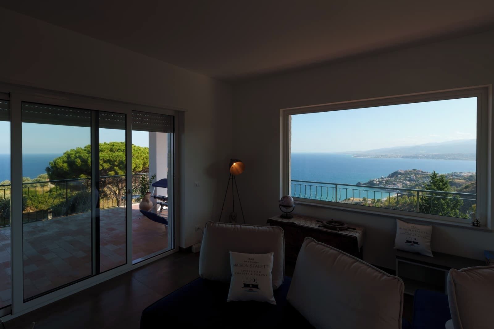 A spacious room with a large window offering a view of the sea and hills. There is a balcony with a table and chairs outside.