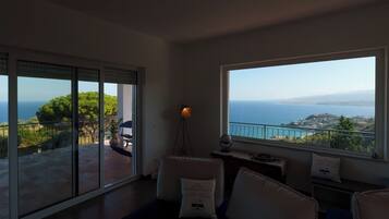 A spacious room with a large window offering a view of the sea and hills. There is a balcony with a table and chairs outside.
