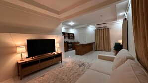 A modern living room with a flat-screen TV, a sofa, and a coffee table. There is a kitchen area in the background with a built-in oven and a sink.