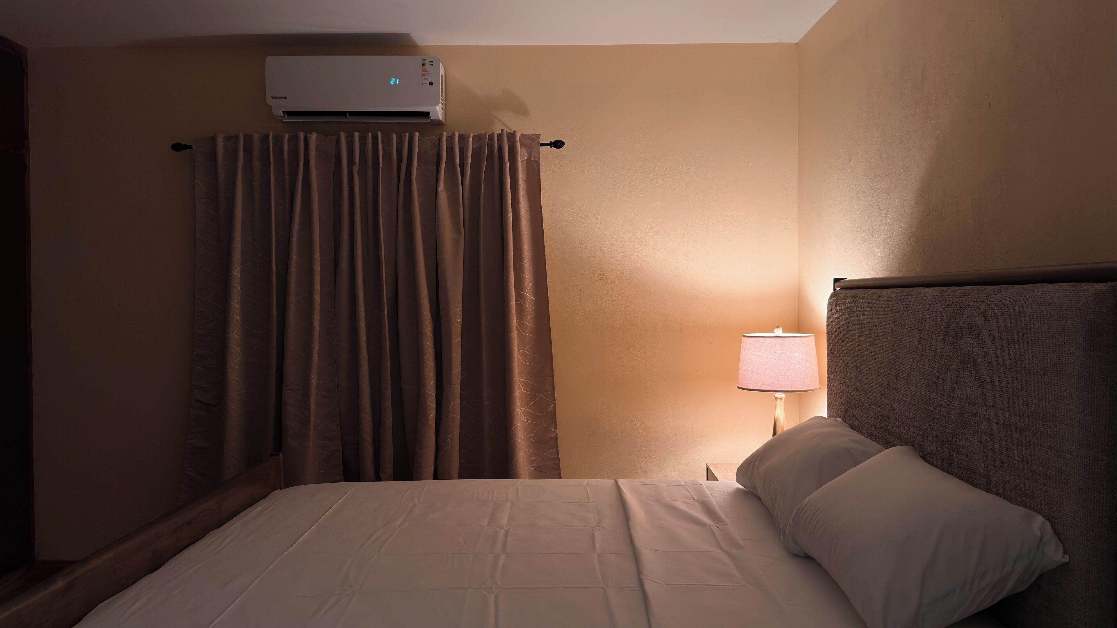 A hotel room with a bed, a lamp, a window with curtains, and an air conditioning unit.