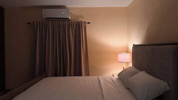 A hotel room with a bed, a lamp, a window with curtains, and an air conditioning unit.