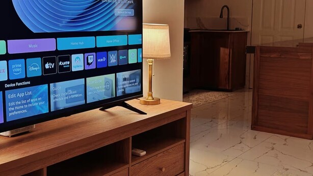 A flat-screen TV on a wooden stand with a lamp beside it, displaying a home hub interface with various app icons.