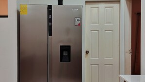 A stainless steel refrigerator with a control panel and a freezer compartment, a white door with a handle, and a wooden cabinet with drawers.