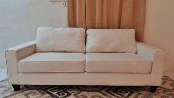 Un sofá beige con dos cojines en una habitación con un tapete estampado y una cortina al fondo.