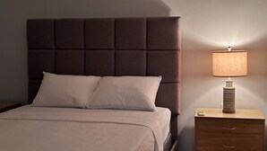 A bed with a padded headboard, two pillows, and a bedside table with a lamp and a remote control.