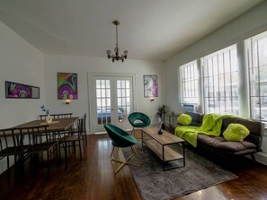 A spacious living room with a wooden dining table, two green chairs, a brown sofa with bright green pillows, and a chandelier.