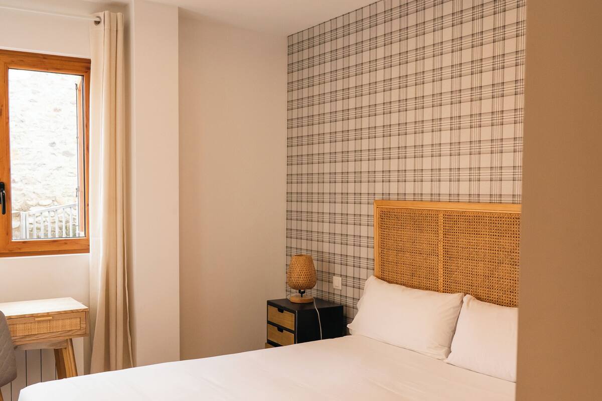 A bedroom with a large bed, a bedside table, a lamp, and a window with curtains.