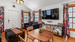 Ein Zimmer mit Esstisch aus Holz, einer schwarzen Couch und einem an der Wand montierten Fernseher.