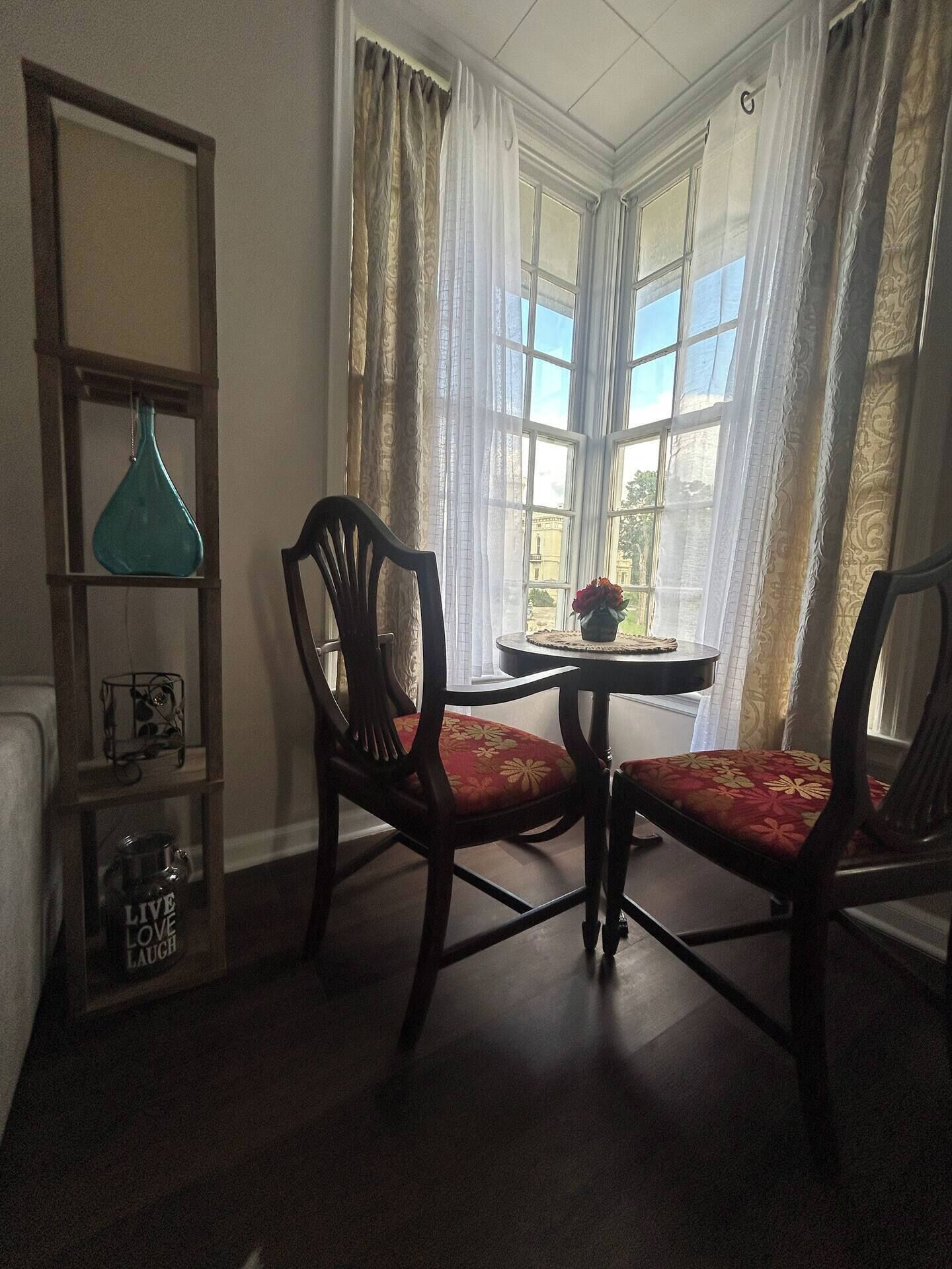 A dining area with two wooden chairs and a small round table, a shelf with decorative items, and a window with curtains.