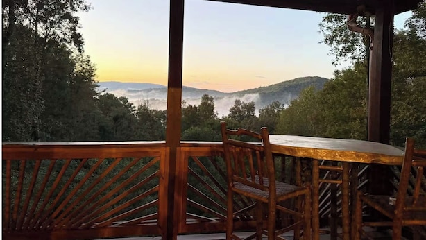 Une terrasse en bois avec une table et des chaises, donnant sur une vallée boisée où les collines sont enveloppées de brume.