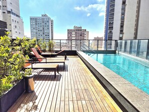 A rooftop pool area with wooden decking, lounge chairs, and a view of surrounding buildings. - Aurora 104 - Estilo e conforto em SP (Sao Paulo)