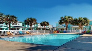 Ein Swimmingpool mit Liegestühlen und Palmen, umgeben von einem weißen Zaun und einer Reihe hellblauer Gebäude.