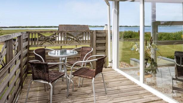 Terraza de madera con sillas de mimbre y una mesa de vidrio, con vista a un lago.