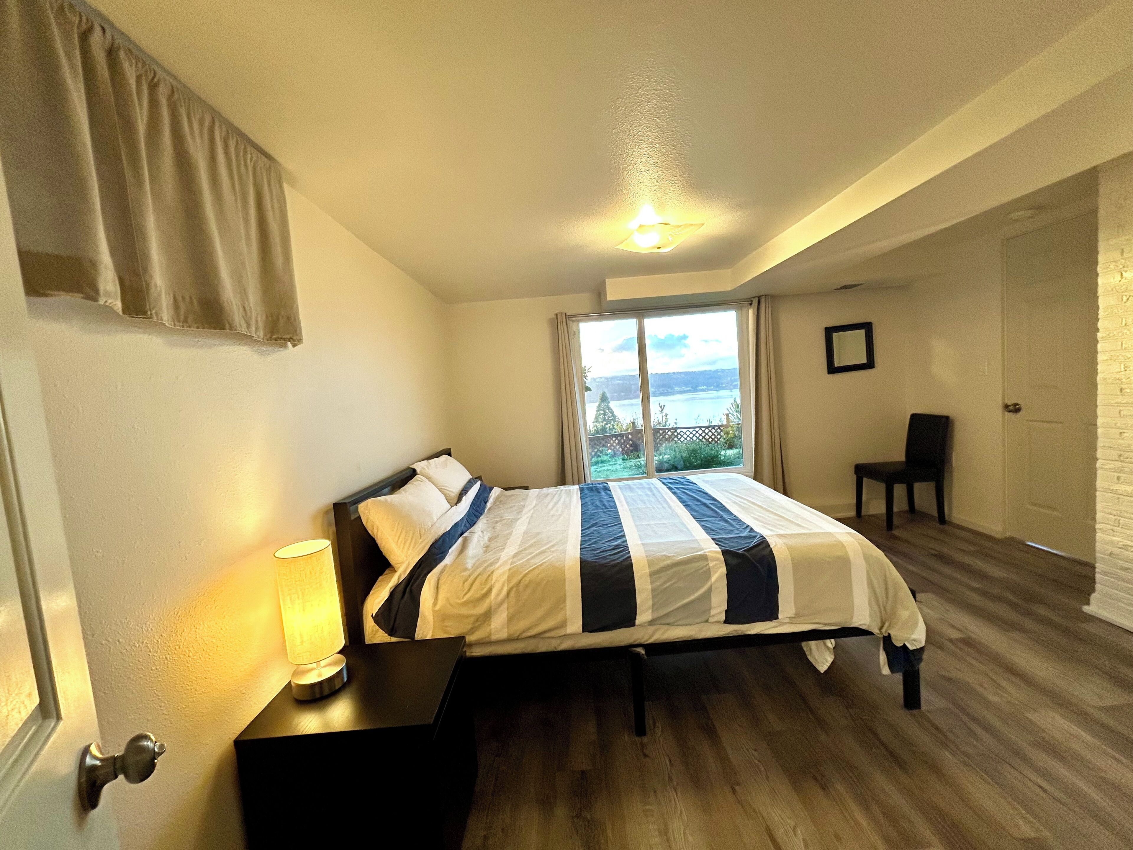 A bedroom with a bed, a nightstand, a chair, a window with a view, and a door.