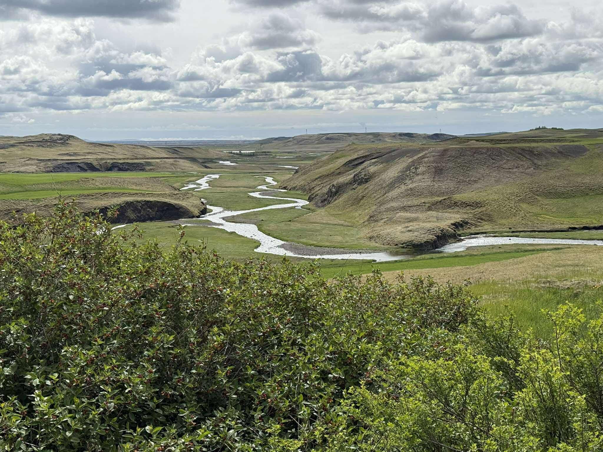 Une rivière serpente à travers une vallée, bordée de collines verdoyantes, sous un ciel nuageux.