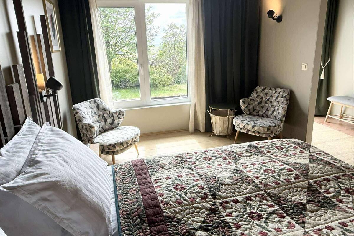Un dormitorio con cama, dos sillones estampados, una ventana con vistas a zonas verdes y una lámpara de noche.