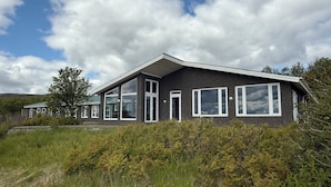 A modern house with large windows and a sloped roof, surrounded by natural vegetation. - Freya Farm (Storinupur)