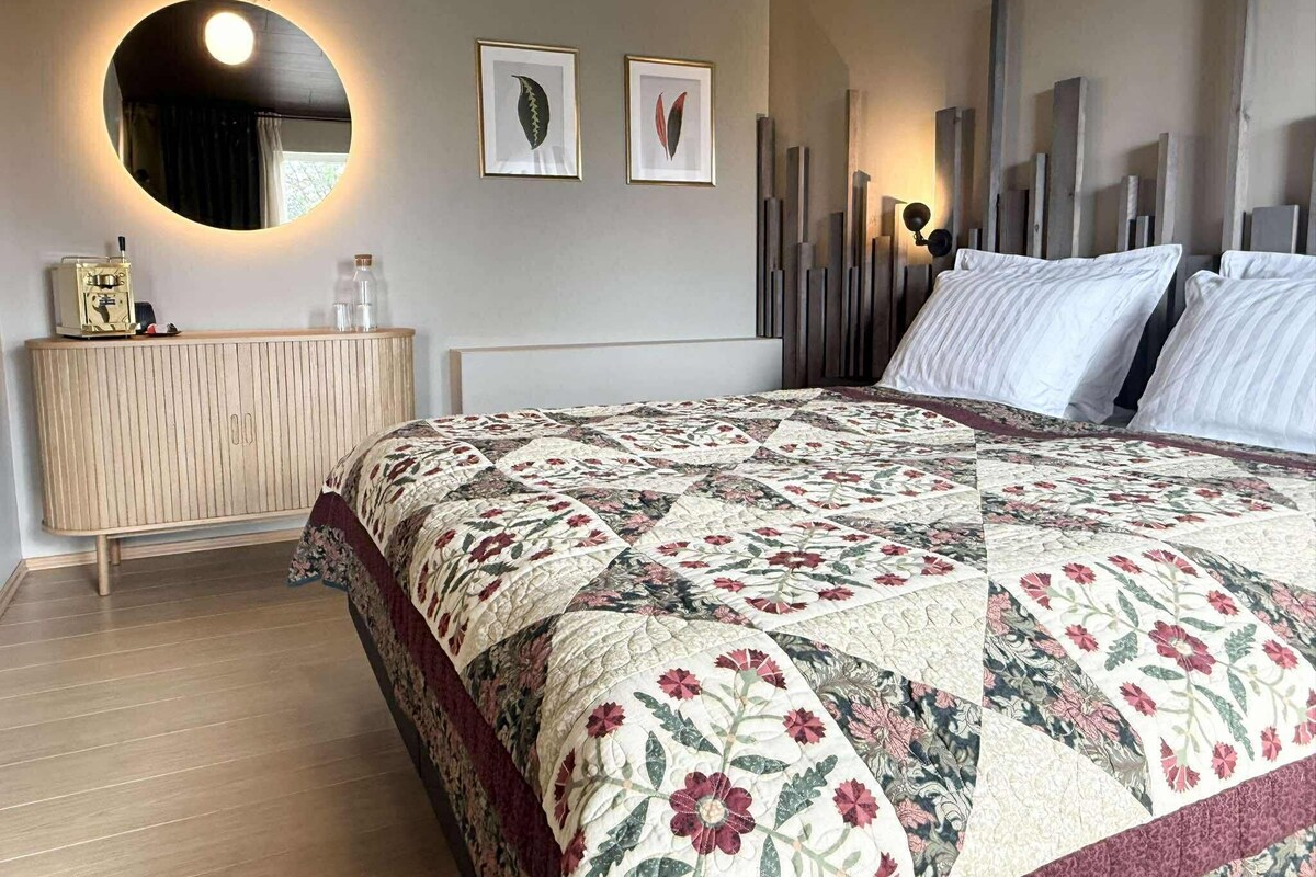 Un dormitorio con una cama grande, un espejo redondo, un aparador y dos láminas botánicas enmarcadas en la pared.