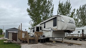 Een Jayco camper met een overdekte veranda en een barbecueplaats, omringd door grind en houten constructies.