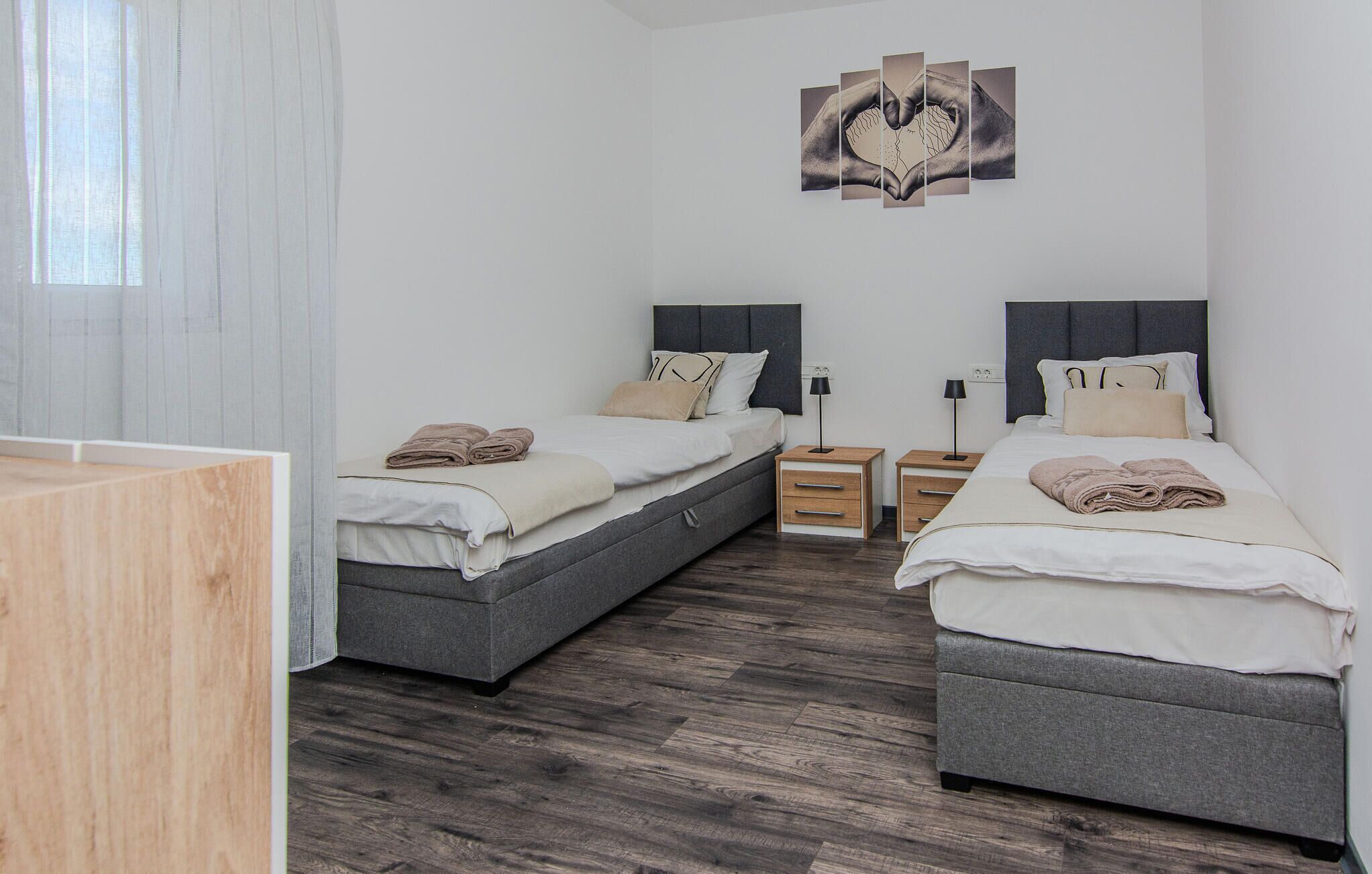 A modern hotel room with two beds, a wooden nightstand, a desk, and a large window with sheer curtains.