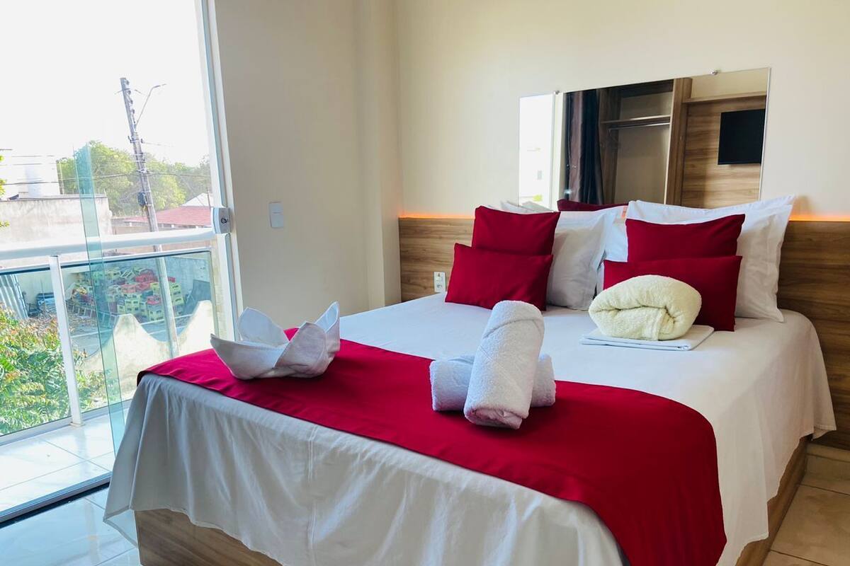 A hotel room with a large bed, red and white bedding, a wooden headboard, and a large window offering a view of buildings and greenery.