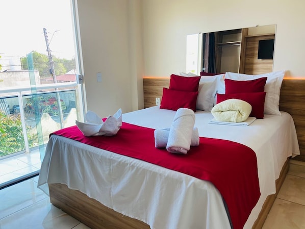 A hotel room with a large bed, red and white bedding, a wooden headboard, and a large window offering a view of buildings and greenery. - Hotel Camocim (Camocim)