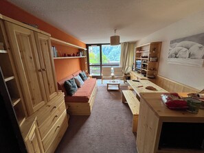 Ein gemütliches Zimmer mit einer Holzcouch, einem Esstisch und einem Fernseher. Regale mit diversen Gegenständen und ein Fenster mit Blick auf Grünflächen.