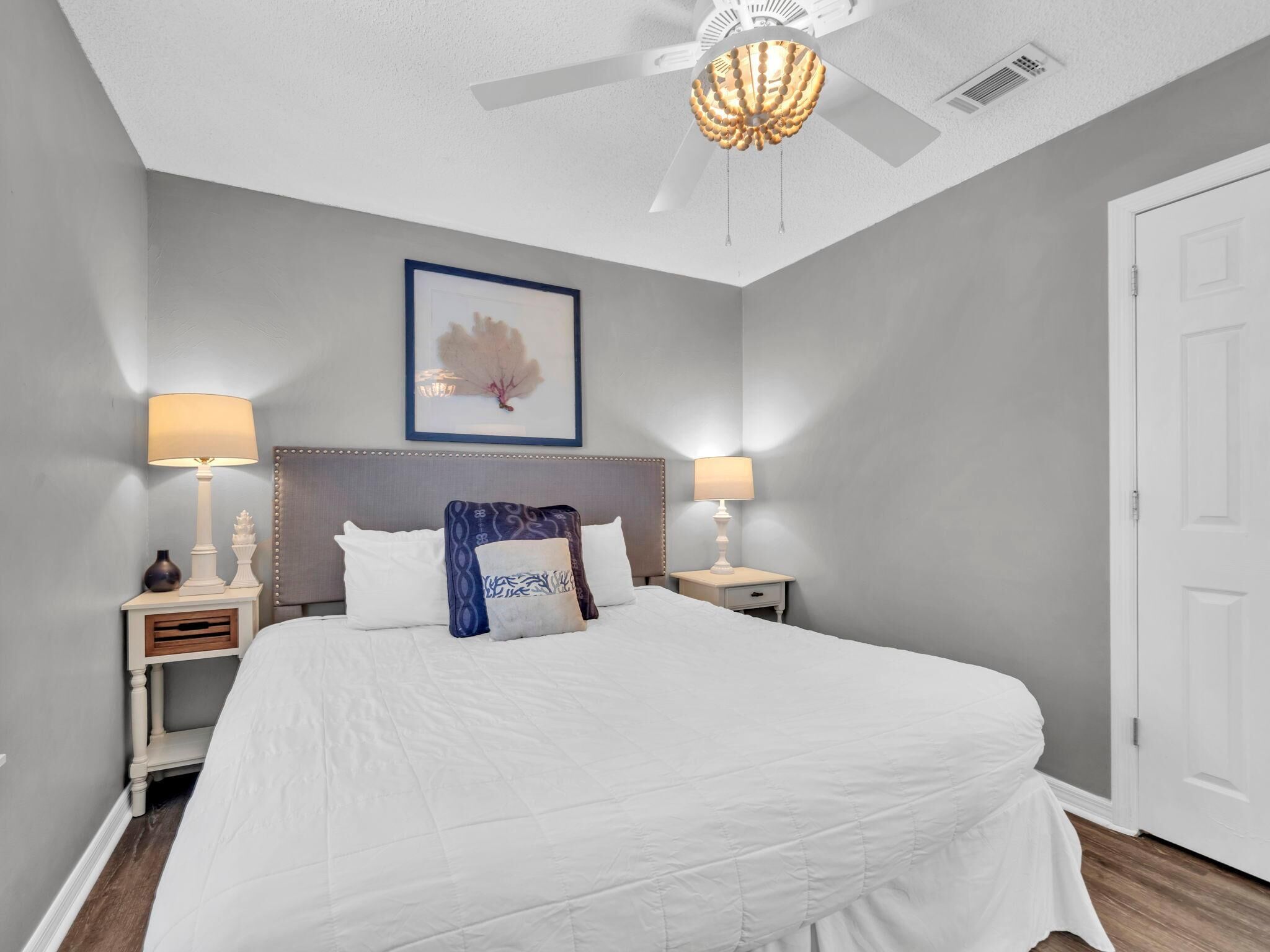 A bedroom with a large bed, two bedside tables with lamps, a ceiling fan, and a framed artwork on the wall.