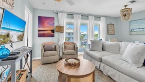 Ein geräumiges Wohnzimmer mit einem weißen Sofa, zwei Sesseln, einem runden Couchtisch und einem Deckenventilator. Große Fenster mit weißen Vorhängen, ein Strandgemälde und ein gerahmter Spruch schmücken die Wände.
