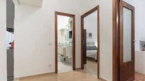 A hallway leading to a bedroom with a bed, a bathroom with a sink and mirror, and a kitchen area visible through an open door. - The little house in the hills - Appio/Tuscolano (Roma)
