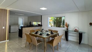 Un comedor amplio con una mesa de vidrio y sillas de mimbre, una cocina al fondo, y un ventanal con vistas a la vegetación.