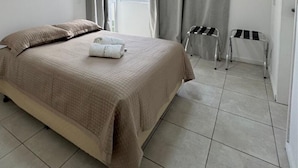 Un dormitorio con cama, ventana, cortinas, radiador y un taburete pequeño.