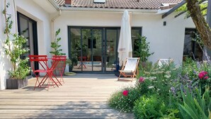 En træterrasse med udendørs møbler, en glasdør og en have med forskellige planter og blomster.