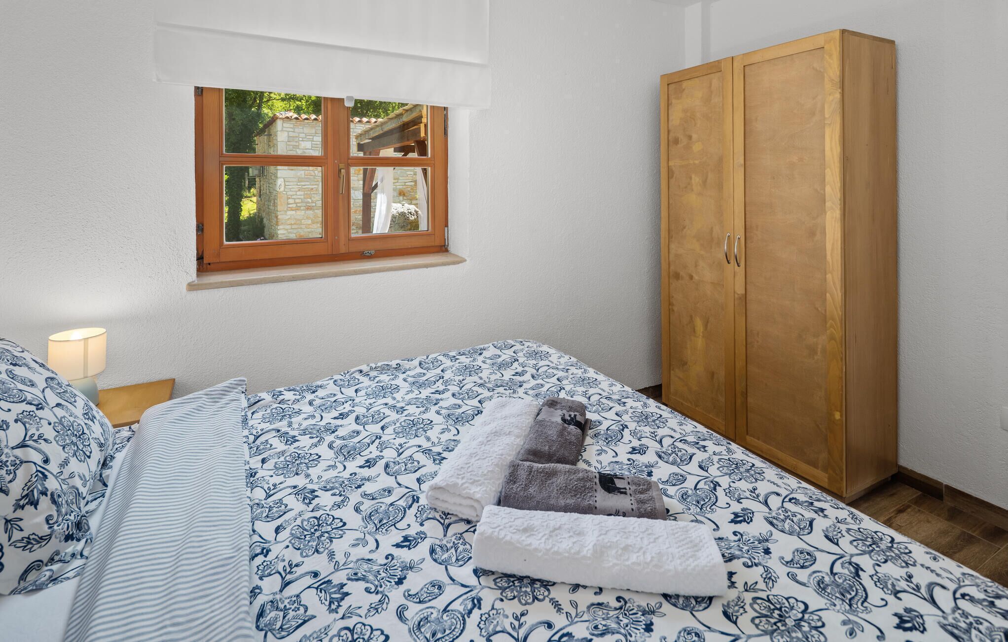 A bedroom with a bed, a wooden wardrobe, a window, and a nightstand with a lamp.