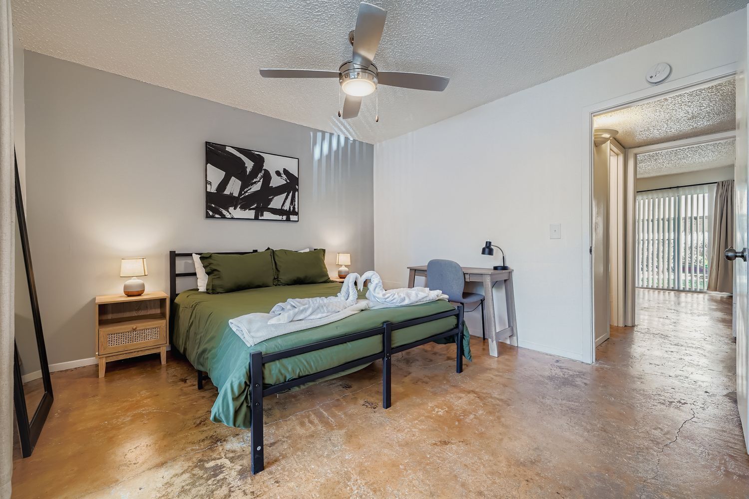 A bedroom with a bed, a desk, a chair, a ceiling fan, and a painting on the wall.