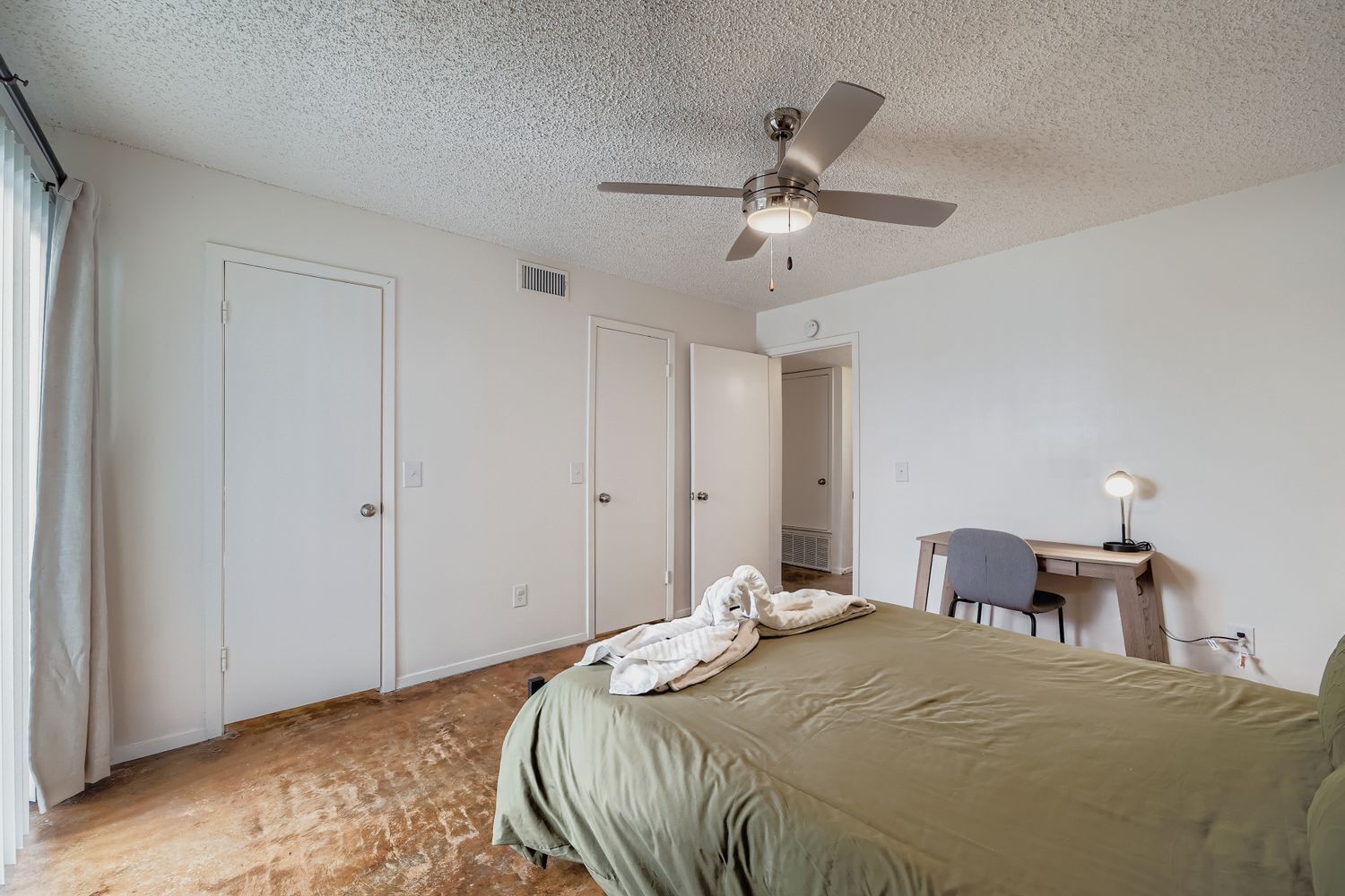 Une chambre à coucher comprenant un lit, un bureau, une chaise, un ventilateur de plafond et une porte.