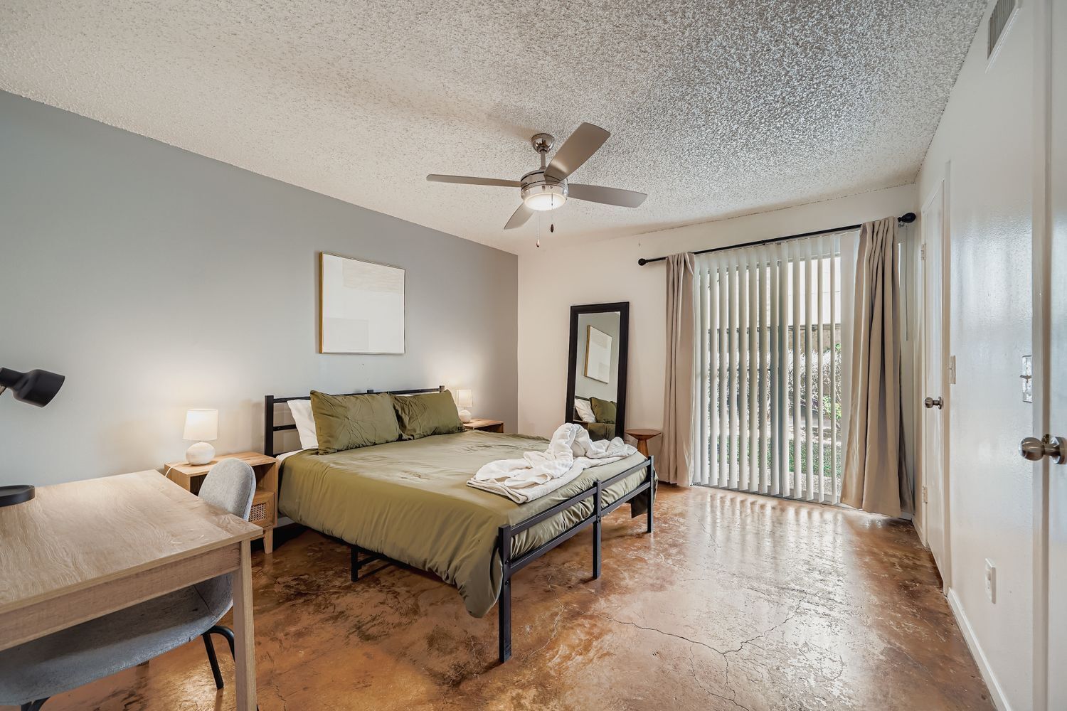 A bedroom with a bed, a desk, a ceiling fan, a window with curtains, and a door.