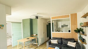 A modern living room with a wooden coffee table, a grey sofa with 'BEACH' pillows, a dining area with a wooden table and chairs, and a kitchen visible through an open door.