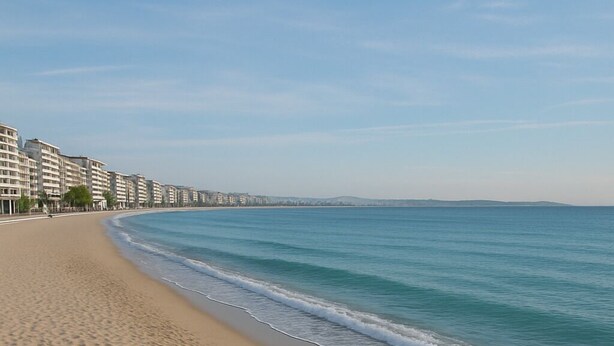 A sandy beach with a clear blue sea and a row of buildings in the background.