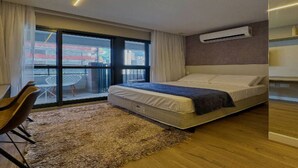 Een moderne hotelkamer met een bed, een klein bureau, een stoel, een groot raam met uitzicht op gebouwen, en een airconditioner aan de muur.