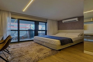 Ein modernes Hotelzimmer mit Bett, kleinem Schreibtisch, Stuhl, großem Fenster mit Blick auf Gebäude und einer Wandklimaanlage.
