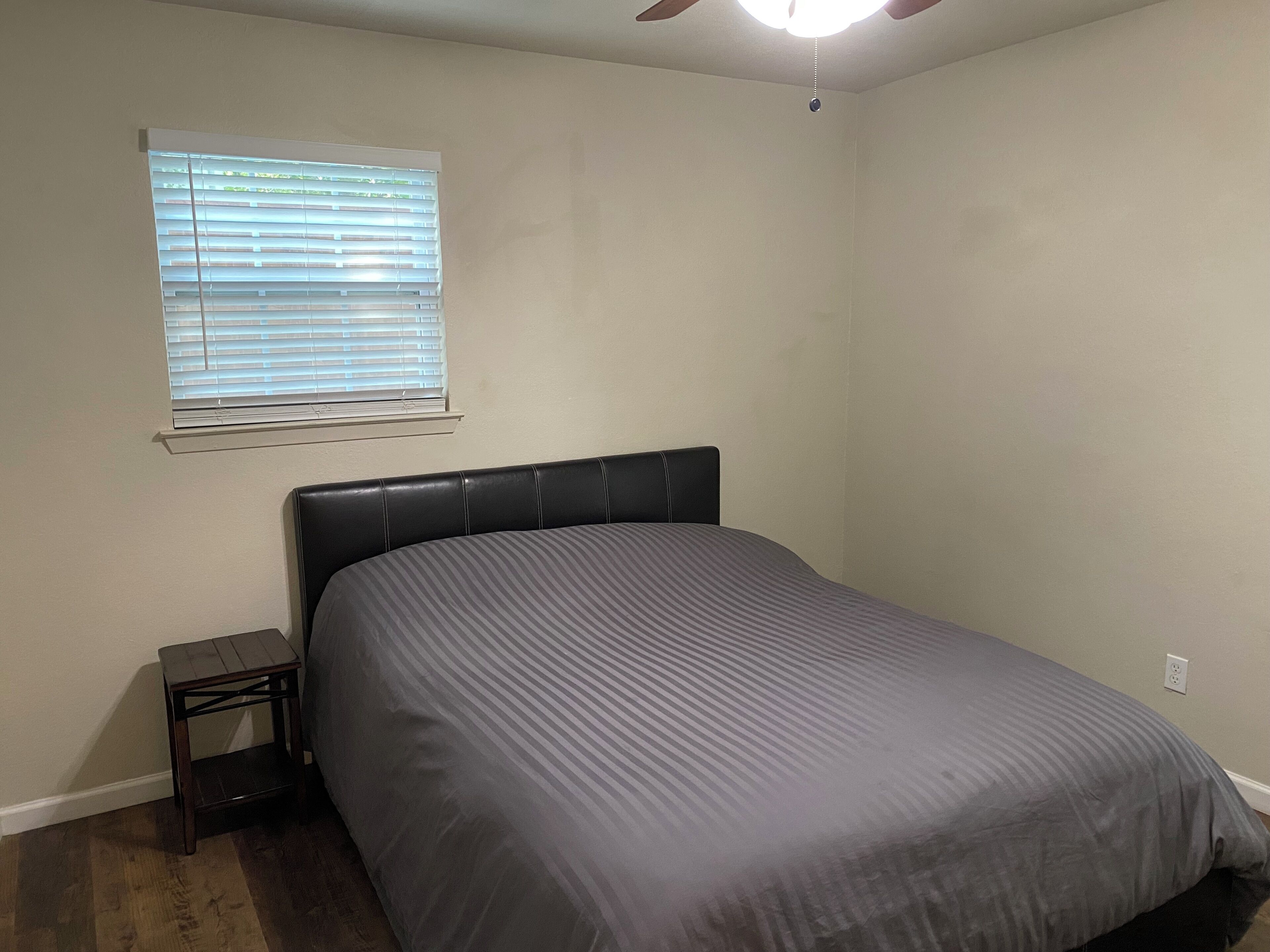 A bedroom with a bed, a nightstand, a window with blinds, and a ceiling fan.