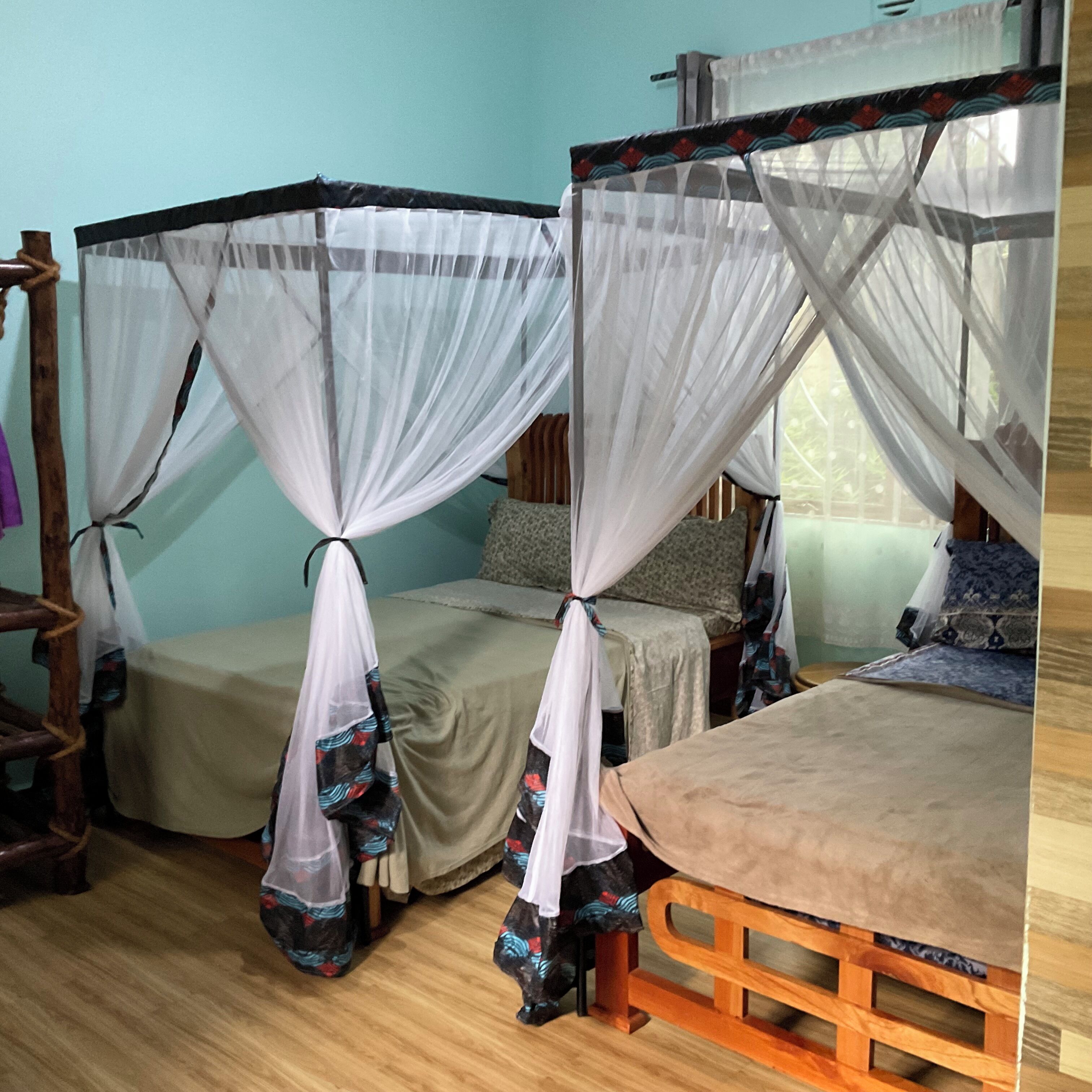 A room with two single beds, each with a canopy bed frame and a mosquito net. There is a wooden table in the foreground.