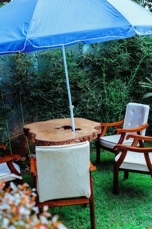 Une table ronde en bois, accompagnée d’une chaise en bois avec un coussin blanc et d’une autre chaise en bois également dotée d’un coussin blanc. Un parasol offre de l’ombre au-dessus de la table.